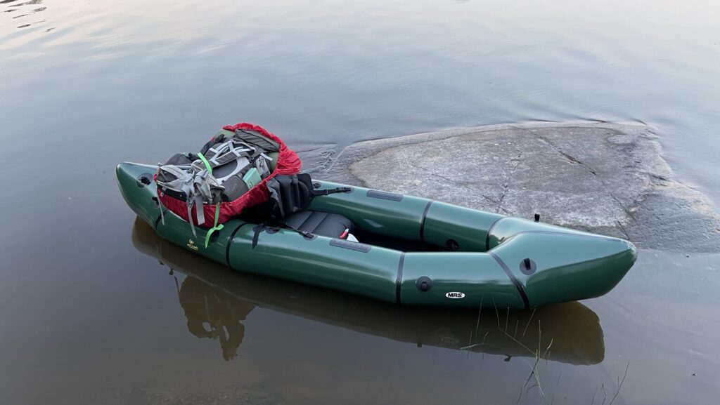 Vihreä packraft tyynessä rantavedessä kallion vieressä kesäiltana, rinkka kyydissä valmiina lähtöön